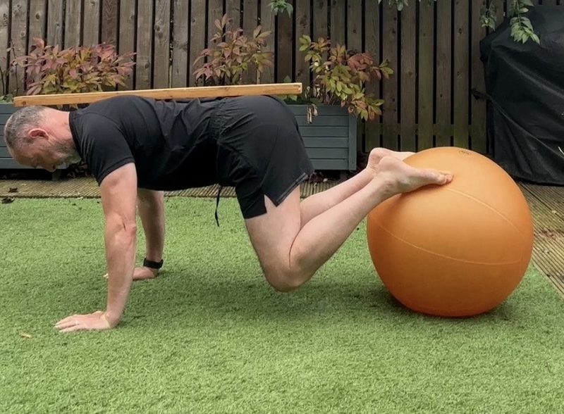 Stephen in a prone jackknife with shins on a Swiss ball, knees drawn in, a stick resting along his spine