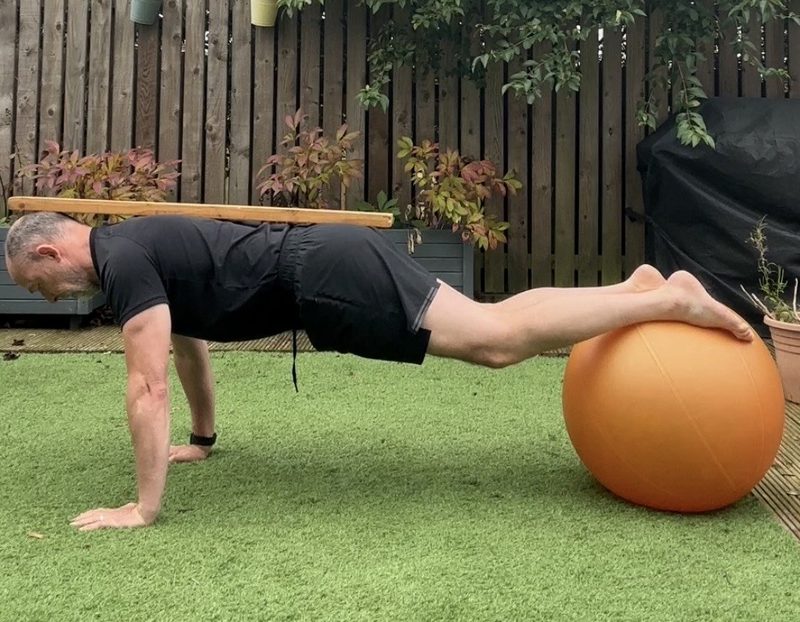 Stephen in a plank with hands on the floor and feet on a Swiss ball, a stick resting along his spine