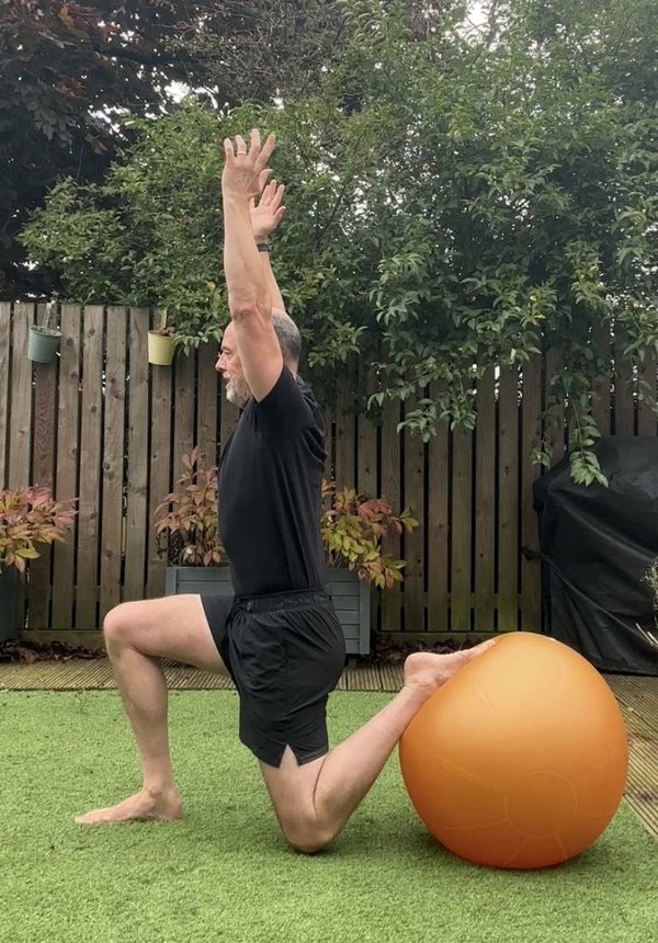 Stephen at the bottom of a Bulgarian split squat, back foot on a Swiss ball, back knee stopping just short of the floor, arms overhead