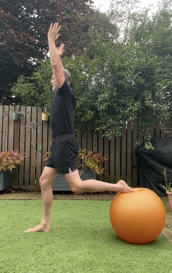 Stephen at the top of a Bulgarian split squat, back foot on a Swiss ball, arms overhead, side view