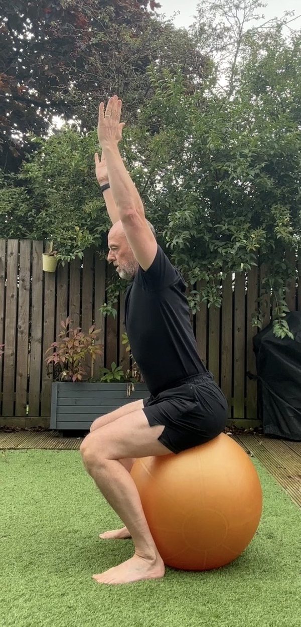 Stephen seated at the bottom of a sit-to-stand on a Swiss ball, arms overhead, side view