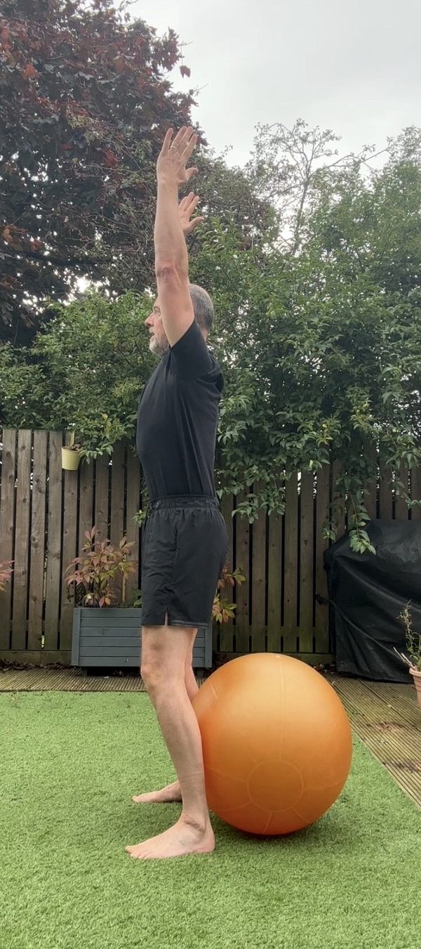 Stephen standing tall at the top of a sit-to-stand in front of a Swiss ball, arms overhead, side view