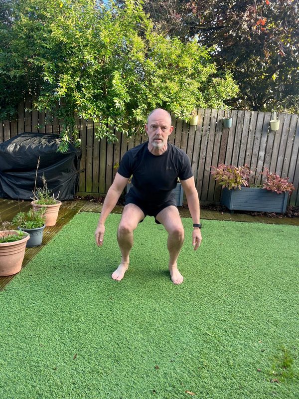 Stephen in a bodyweight squat, front view, arms by his sides, knees over his feet and heels on the ground